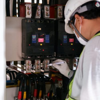 Technician wearing safety gear inspecting electrical panels in an industrial setting.